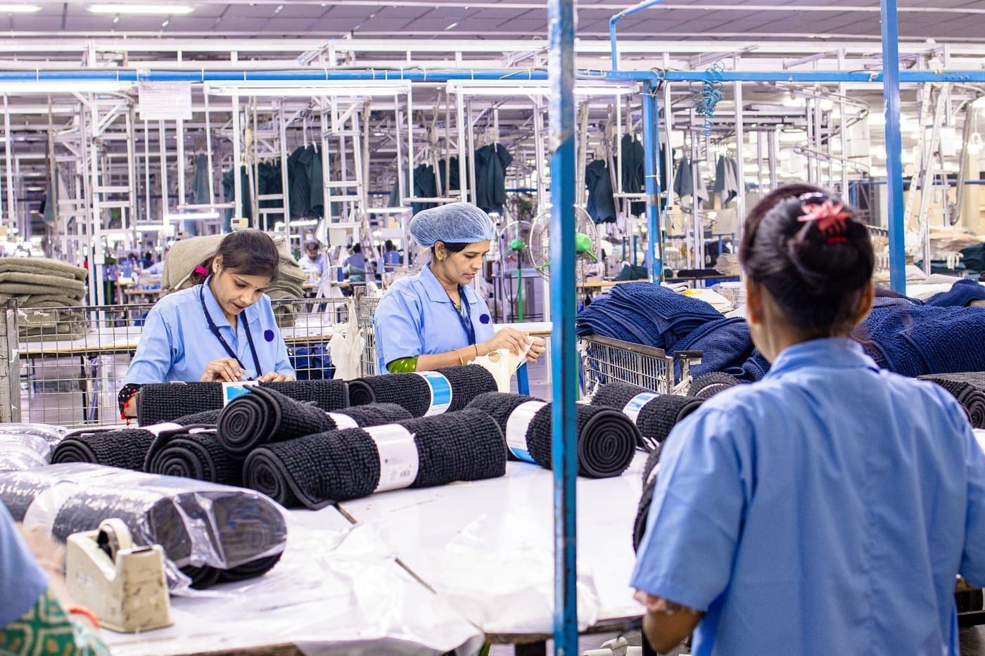 Workers in blue uniforms packing rolled mats at long tables on a large factory floor