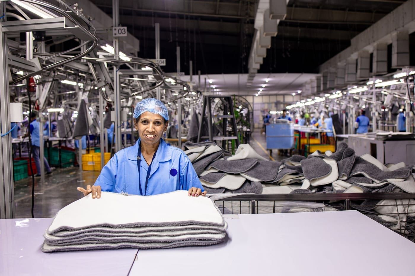 A checker in a hairnet and blue uniform with a stack of finished pieces at a checking table on a textile factory floor