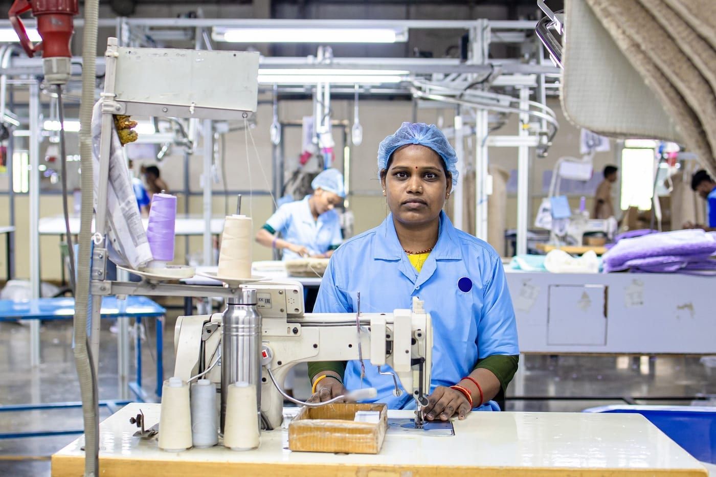 A worker in a blue uniform and hairnet at an industrial sewing machine on a garment factory floor