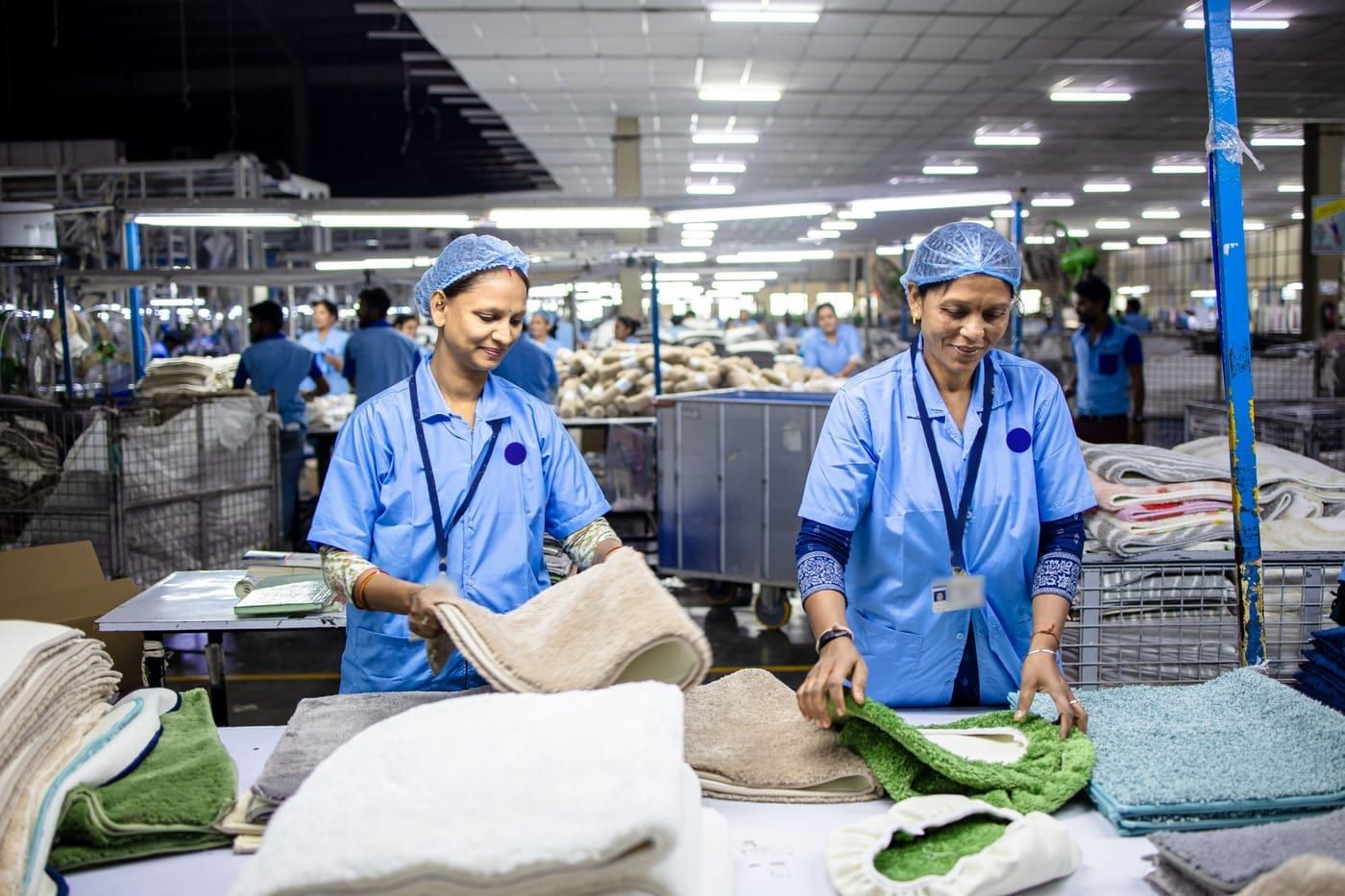Two packers in hairnets checking and refolding mats at a table on a busy factory floor