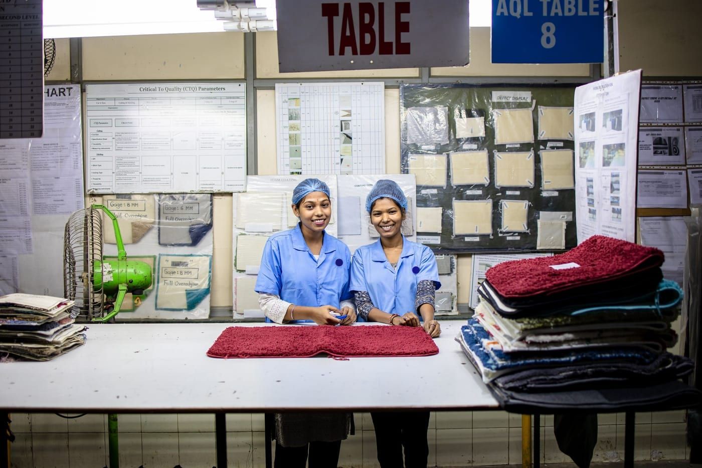 Two inspectors at an inspection table with finished mats, with quality charts and a defect display board behind them