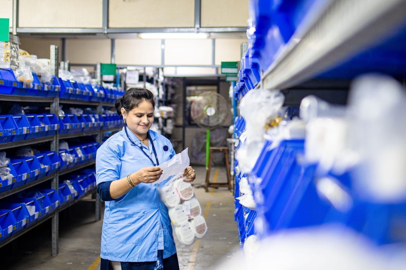 A stores worker in a blue uniform checking a slip against a bag of parts in an aisle of blue bins