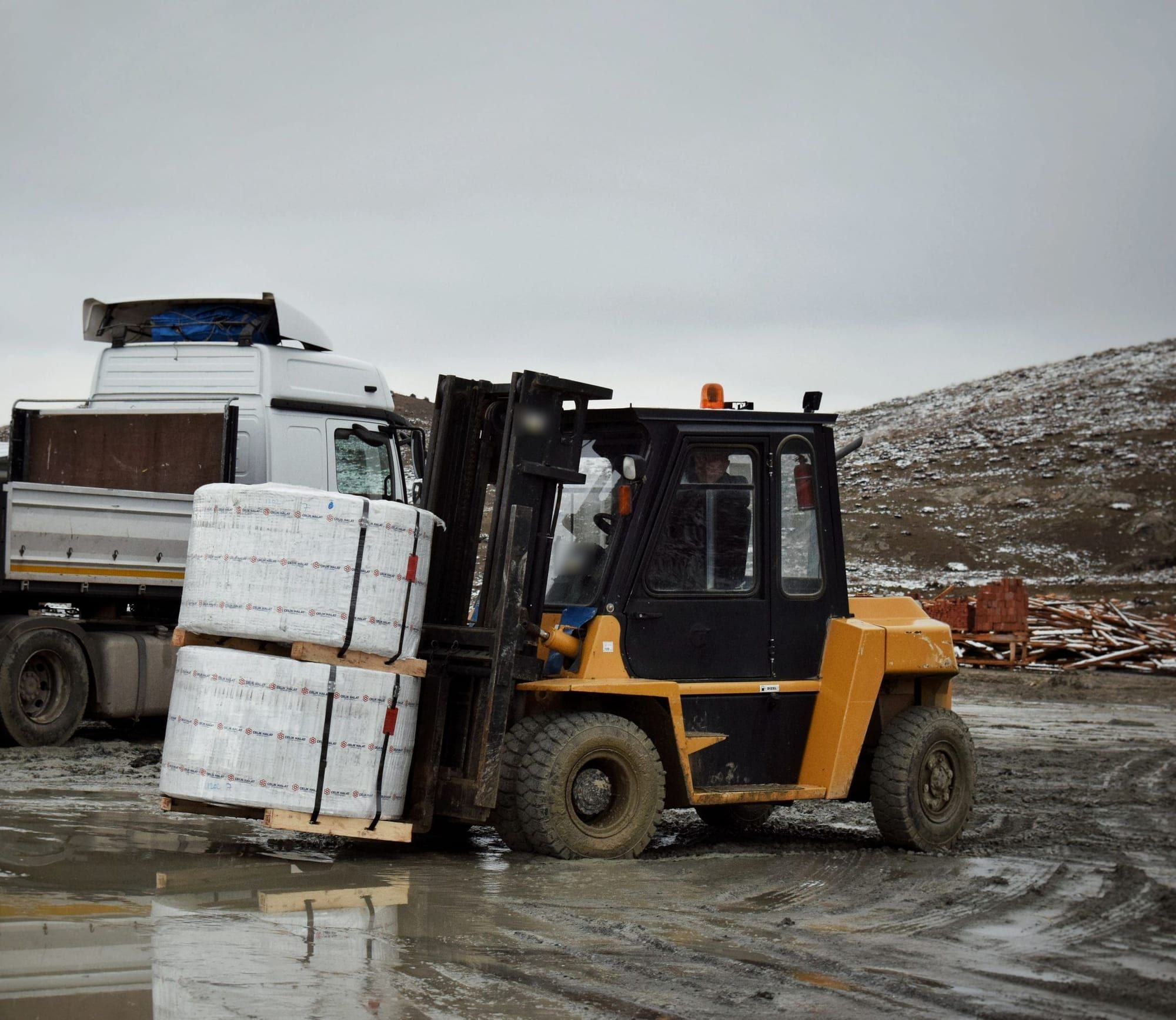 A forklift carrying stacked material across a yard, with a truck behind it